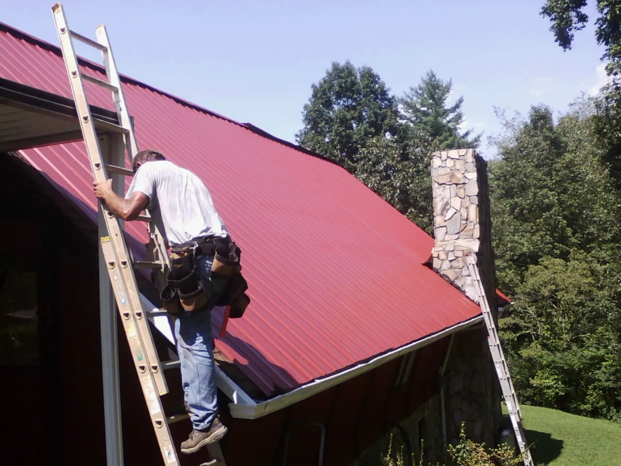 Roofer on a ladder beside a red metal roof and stone chimney — Soffit & Fascia Repair by Roofing Brothers in Sabina